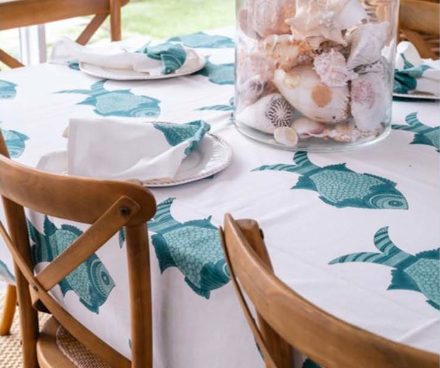 A white tablecloth with a teal fish pattern is set on a dining table, styled with wooden chairs, white napkins, and a large glass vase filled with seashells.